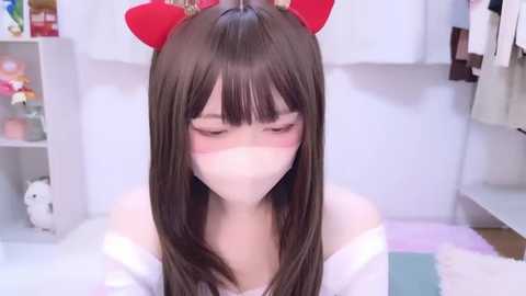 Video of a young woman with fair skin, wearing red cat ears, white mask, and white off-shoulder top, sitting on a plush carpet in a bright, cluttered room with shelves filled with toys and clothes.