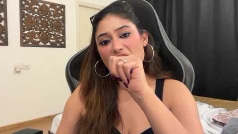 Video of a young South Asian woman with long brown hair, wearing hoop earrings and a black tank top, sitting in a gaming chair. Background features black curtains, a white wall, and ornate wooden art.