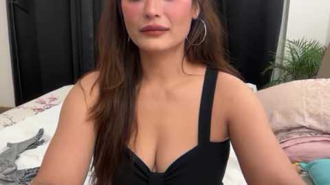 A video of a young woman with long brown hair, wearing a black bra, sitting on a bed with floral sheets and a black curtain in the background.