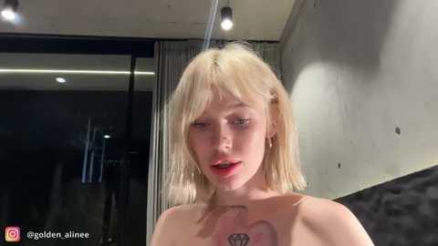Video of a topless, blonde woman with fair skin and a tattoo on her chest, standing in a modern, dimly lit room with concrete walls and recessed lighting.