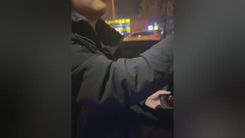 Video of a dark-haired woman with a large black coat, holding a cellphone, standing in front of a car at night. Background shows blurred yellow neon signs and dim streetlights.