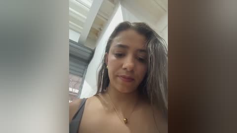 Video of a young woman with medium skin tone, long dark hair, wearing a black top, standing indoors near a window.