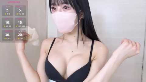 A video of an East Asian woman with long black hair and fair skin, wearing a pink face mask and a black bra, flexing her arms in a bathroom with a visible thermometer.