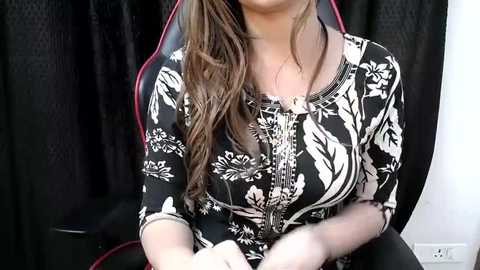 Video of a woman with long, wavy brown hair, wearing a black floral-patterned dress with a zipper, seated indoors against a black curtain background.