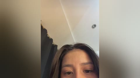 Video of a young woman with light brown hair, looking directly at the camera. The background shows a ceiling with a visible crack and a light fixture. Her eyes are visible, and her expression is neutral.