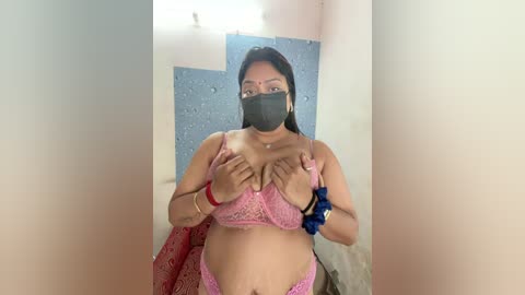 A video of a South Asian woman with medium-dark skin tone, wearing a pink lace bra, a black face mask, and blue scrunchies, standing in a narrow, brightly lit hallway.