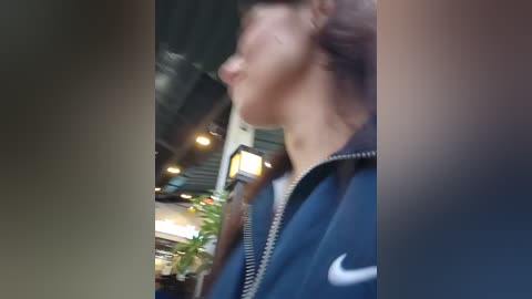 Video of a person in profile wearing a blue jacket with a white Nike logo, standing in a modern indoor space with exposed ceiling beams, potted plants, and ambient lighting.