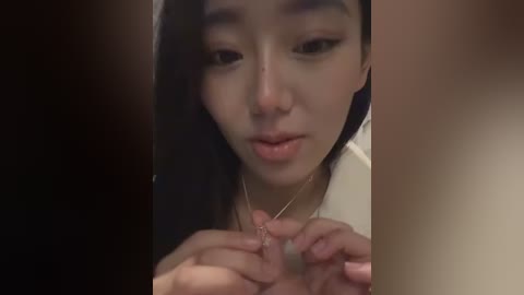 Video of an Asian woman with long black hair, focused expression, wearing a white top, delicately adjusting her gold necklace in front of a blurred background.