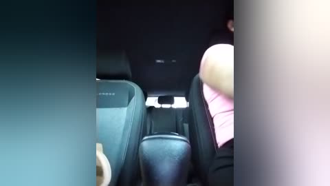 A video of a car interior showing a person in a pink shirt sitting on the right side, facing forward. The seats are black with beige accents, and the lighting is dim.