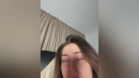 Video of a young woman with long, straight brown hair, wearing a beige top, partially obscured by a beige curtain. Her face is blurred, with a neutral expression.