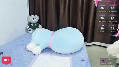 Video of a plush, light blue teddy bear lying on a bed with a green jersey, gray teddy bear, and brown curtains in the background.