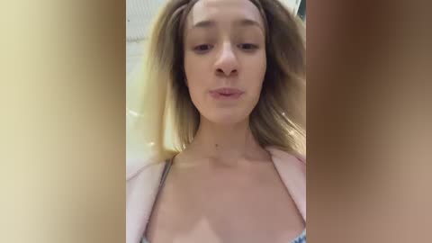 A video of a young woman with light skin, medium-length brown hair, and a neutral expression, wearing a light-colored top, taken from a close-up angle.