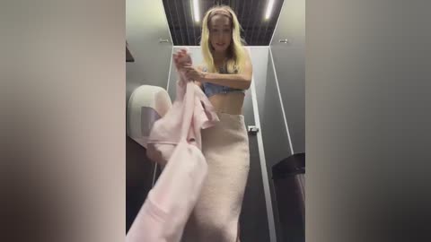 Video of a blonde woman in a bathroom, wearing a blue bra and beige towel, holding a pink towel, with a modern, minimalist design.