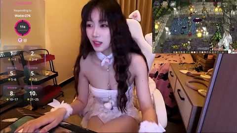 Video of an Asian woman in a white bunny costume, sitting at a cluttered desk with a gaming monitor displaying a virtual world.