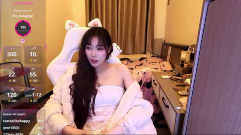 Video of an East Asian woman in a white dress with long black hair, sitting on a bed in a bedroom. She has a playful expression, and the room has a cozy, warm atmosphere.