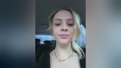 A video of a young, fair-skinned woman with blonde hair, wearing a black jacket, and a delicate necklace, sitting in a dimly lit car. The background is blurry, featuring indistinct shapes.