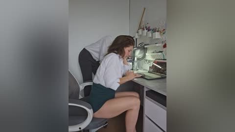 Video of a young woman with long brown hair, wearing a white blouse and green skirt, sitting at a cluttered kitchen counter, looking at a laptop.