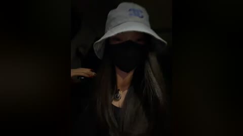 Video of a woman with long dark hair, wearing a white bucket hat, black face mask, and a gray jacket, with a necklace visible.