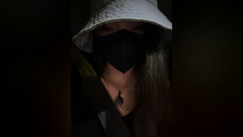 A close-up video of a woman in shadow, wearing a white bucket hat, black face mask, and dark coat, with a pendant necklace, in a dimly lit, dark background.