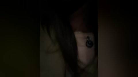 A dimly lit video shows a close-up of a person with long, dark hair partially obscuring their face. A small, black earring is visible on their ear. The background is dark and indistinct.