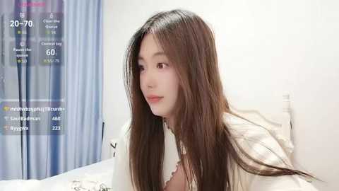 A video of a young East Asian woman with long, straight brown hair, wearing a white top, sitting on a bed. A digital clock and weather display is visible in the background.