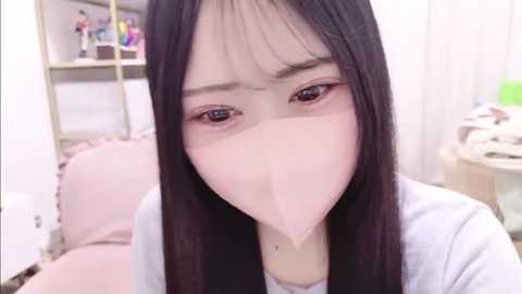A video of an East Asian woman with long black hair, wearing a pink surgical mask, looking concerned in a cluttered room with shelves filled with books and stationery.