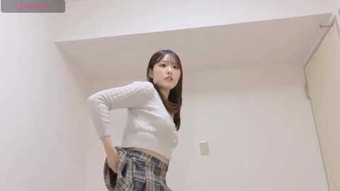Video of a young East Asian woman with long brown hair, wearing a white knit sweater and a plaid skirt, standing confidently in a minimalist white room with a light switch on the wall.