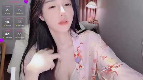 Video of a young Asian woman with long black hair, fair skin, and a pink floral robe, in a bedroom with a bed, curtains, and a lamp.