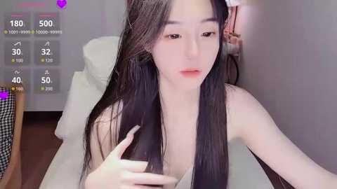 Video of an East Asian woman with long black hair, fair skin, and slender physique, sitting topless on a bed, with a video overlay showing viewers' interactions.