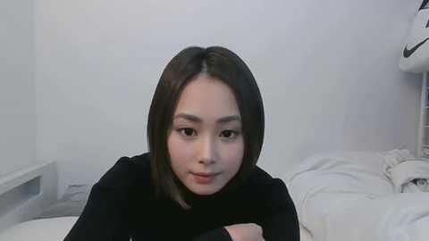 Video of a young East Asian woman with straight, shoulder-length black hair, wearing a black sweater, lying on a white bed in a minimalist, white room with a gray floor.