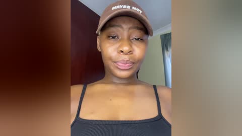 Video of a young Black woman with medium skin tone, wearing a black tank top and brown \"Maybe Not\" cap, making a pouty face. Background features a red wall and a blurred doorway.