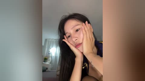 Video of an Asian woman with long black hair, resting her face on her hands, in a dimly lit room with a white curtain and a lamp.