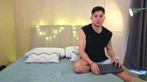 Video of a muscular, tattooed, short-haired man in a black sleeveless shirt and shorts, sitting cross-legged on a bed with white pillows and a gray headboard, typing on a laptop in a cozy, softly lit bedroom.