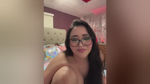 Video of a fair-skinned woman with long black hair, wearing glasses, and a pink shirt, sitting on a bed with a tufted headboard and a colorful bedspread.