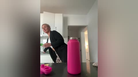 A video of a woman with short blonde hair, wearing a black robe, bending over a kitchen counter with a pink water bottle and a green and white bowl, in a modern, minimalistic kitchen.