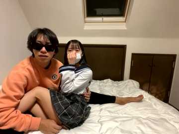 Video of two Asian women, one with short dark hair, wearing a white shirt and plaid skirt, and the other with long hair, a white shirt, and plaid skirt, lying on a bed, both wearing masks, in a dimly lit room with a skylight.