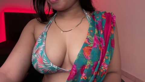 Media: Video of a woman with medium skin tone and dark hair, wearing a turquoise bikini with a floral pattern and a vibrant, floral sarong draped over her shoulder. She's indoors with a pink wall and red lighting.