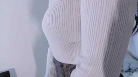 A video of a woman in a white, ribbed sweater and black plaid skirt, standing with her back to the camera, highlighting her slim waist and curvy hips. The background is blurred, showing a plain, light-colored wall.