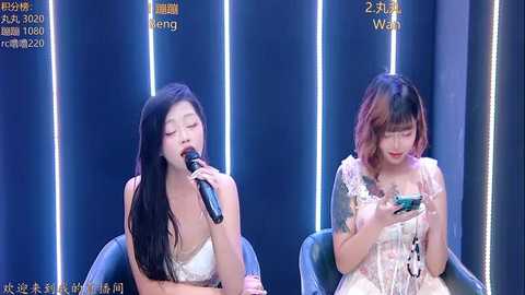 Video of two Asian women singing into microphones against a dark blue, lit-up backdrop; one wears a white dress, the other a floral gown.