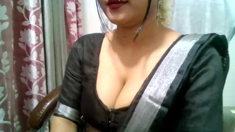 Video of a woman with medium skin tone, wearing a black blouse with a deep neckline, revealing ample cleavage. She has long, wavy blonde hair, and a silver shoulder strap. Background features red and white floral curtains.