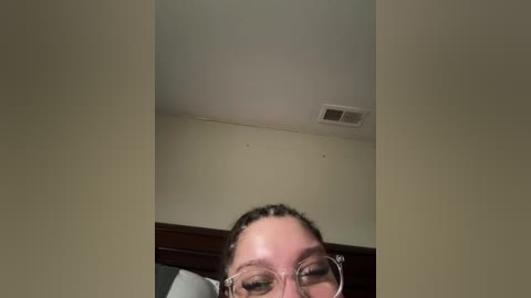 Video of a woman with fair skin, glasses, and dark hair, partially visible from the neck up. She stands in a room with beige walls, a wooden door, and a ceiling light fixture.