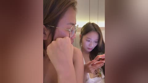 Video of an Asian woman with glasses and long brown hair, holding a smartphone, looking at her reflection in a mirror, while another woman with long dark hair and a white tank top stands behind her.
