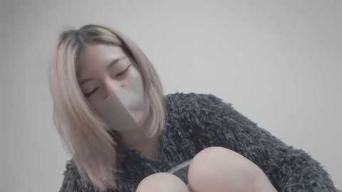 Video of a young Asian woman with shoulder-length blonde hair, wearing a dark gray fuzzy sweater, gagged with white tape, eyes closed, against a plain light gray background.