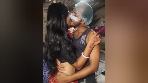 Video of a Black man with a beard wearing a blue face shield, holding a Black woman with long black hair, wearing a black crop top and beige underwear, in a dimly lit bedroom.