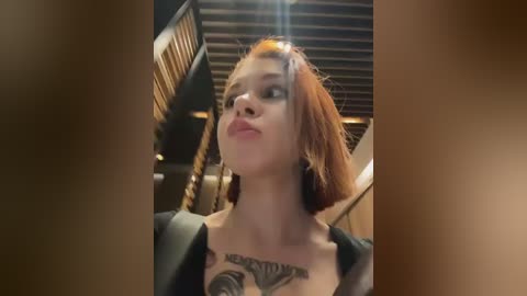 Video of a young woman with fair skin, short red hair, and multiple piercings, wearing a black top that reveals a tattoo on her chest. She stands in a modern, dimly-lit room with wooden slats on the ceiling.