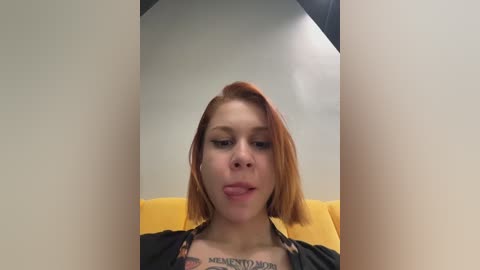 Video of a young, fair-skinned woman with shoulder-length, auburn hair, wearing a black top with a tattoo on her chest. She's sitting on a yellow chair, looking directly at the camera.