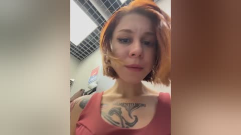 Video of a pale-skinned woman with short, orange hair, wearing a pink tank top, revealing a detailed tattoo on her chest. She's indoors with fluorescent lighting and a grid ceiling.