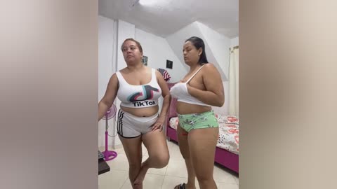 Video of two plus-sized women in revealing TikTok-themed crop tops and shorts, standing in a cluttered bedroom with a bed and pink furniture.