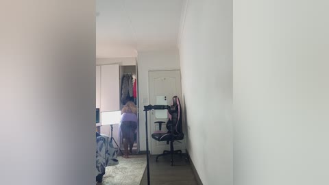 A video of a minimalist bedroom with a mirror showing a woman in a purple dress standing in front of a white door and a black office chair.