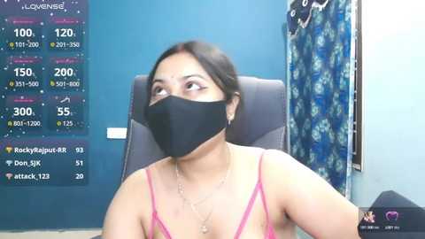 Video of a young Asian woman with medium skin tone, wearing a black face mask, pink spaghetti-strap top, and black necklace, seated in a blue room with a financial chart and blue floral curtain in the background.
